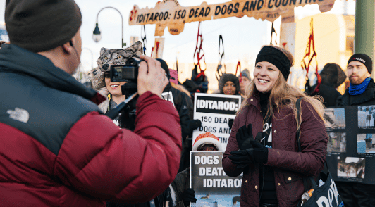 woman doing an interview standing in front of a bunch of protesters at the Iditarod