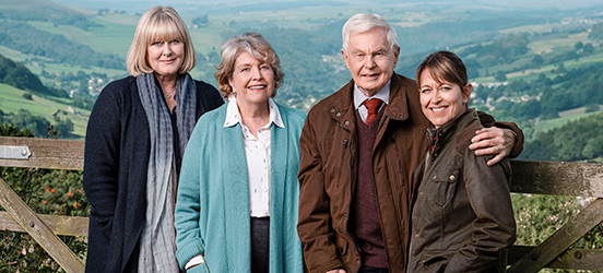 Two older women, an older man, and a middle-aged woman smile as they stand in front of a background of rolling green hills.