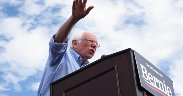 Bernie Sanders at a podium giving a campaign speech against a cloudy blue sky