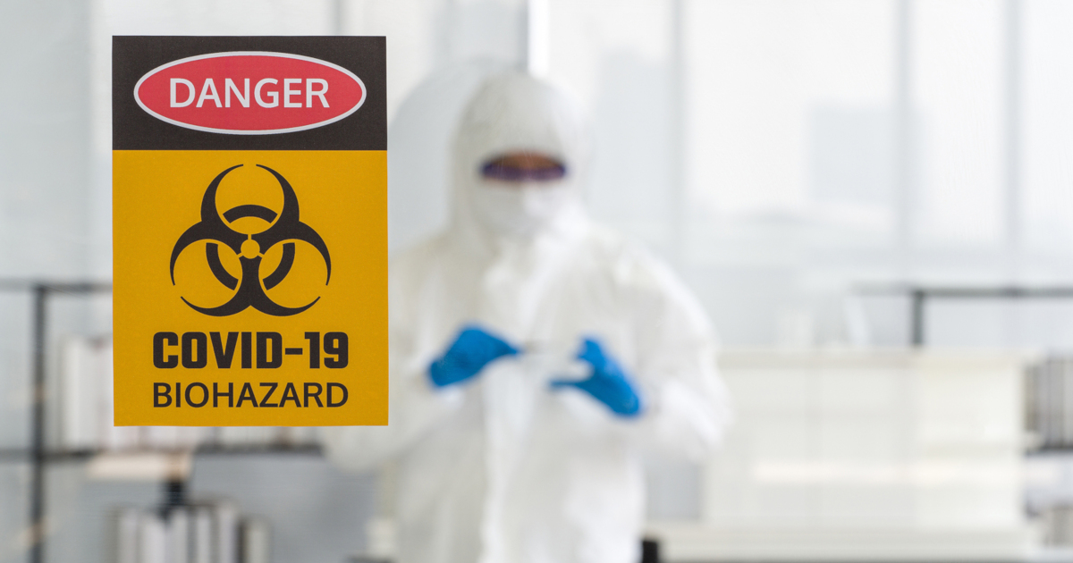 person in a hazmat suit in a laboratory behind a glass door with the warning signs for COVID-19
