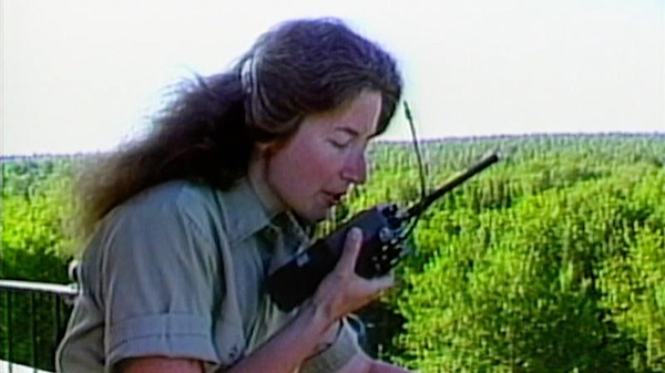 A woman dressed in a collared shirt wears headphones and speaks into a walkie talkie from atop a lookout tower.