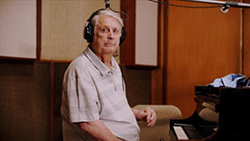 A man wearing headphones and a collared shirt sits inside a musical studio at a piano. 