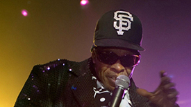 A man wearing a black sequin tuxedo and black baseball cap with white embroidered letters S and F sings into a microphone against a colorful background.