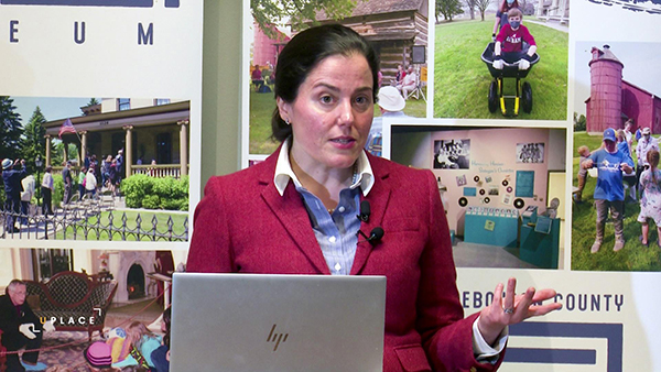 Sarah Anne Carter, a woman with brown hair wearing a red blazer and blue shirt, stands behind a laptop computer giving a lecture.