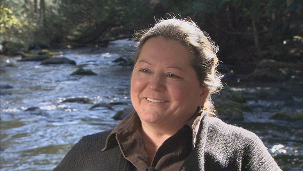 Kim Vele, a female member of the Stockbridge-Munsee Mohican Community, sits in front of the rolling waters of a river.