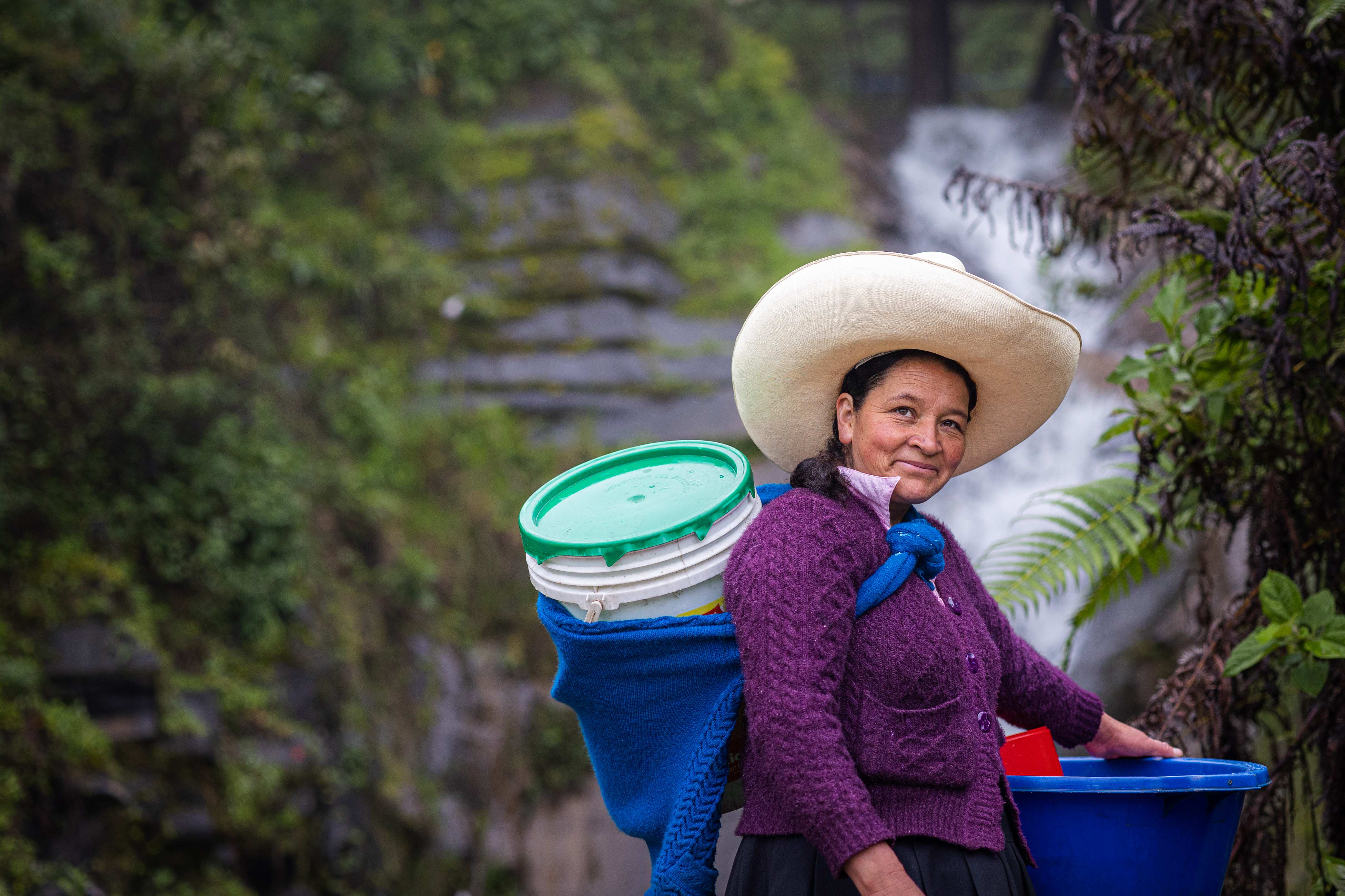 A woman in a hat in front of a waterfall carrying buckets