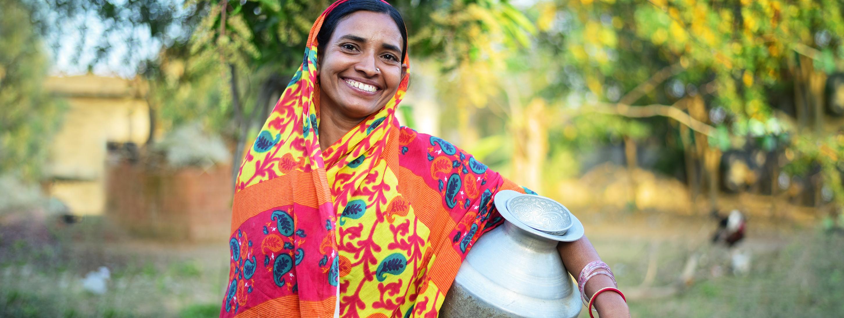 A woman in front of a field smiling and holding a water vessel