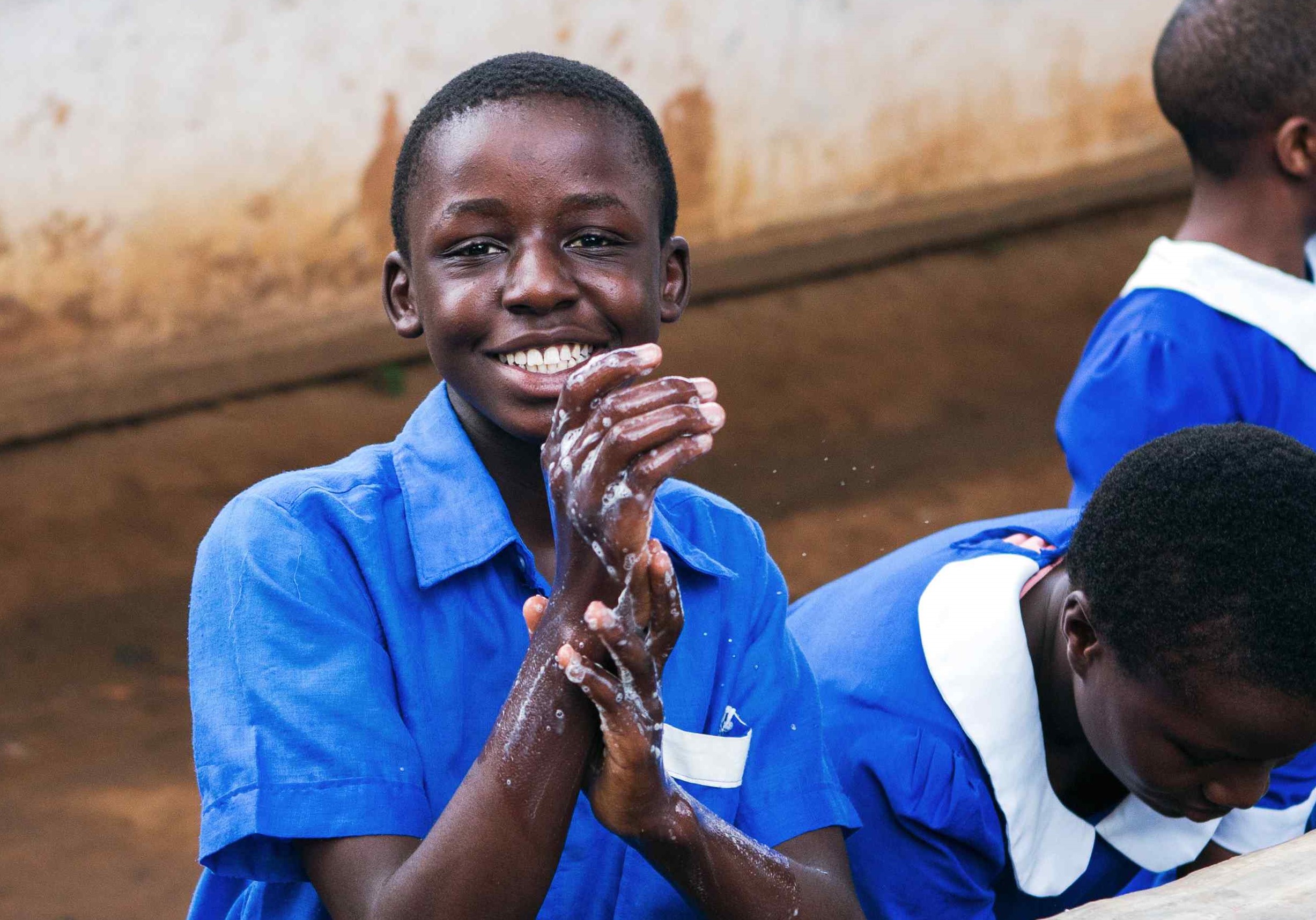 A student in uniform smiling while washing their hands outside at a handwashing station