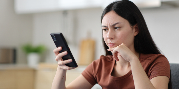 A woman looks at her smartphone with a thoughtful, skeptical expression while sitting at home.