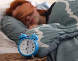Alarm clock showing the time on a bedside table as a person sleeps in the background wearing a sleep mask.