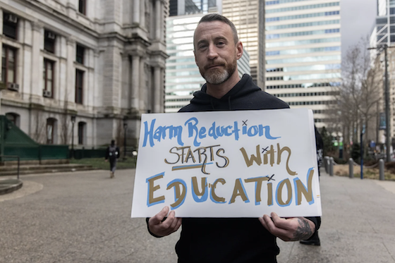 A person stands holding a sign that says, "Harm reduction starts with education."