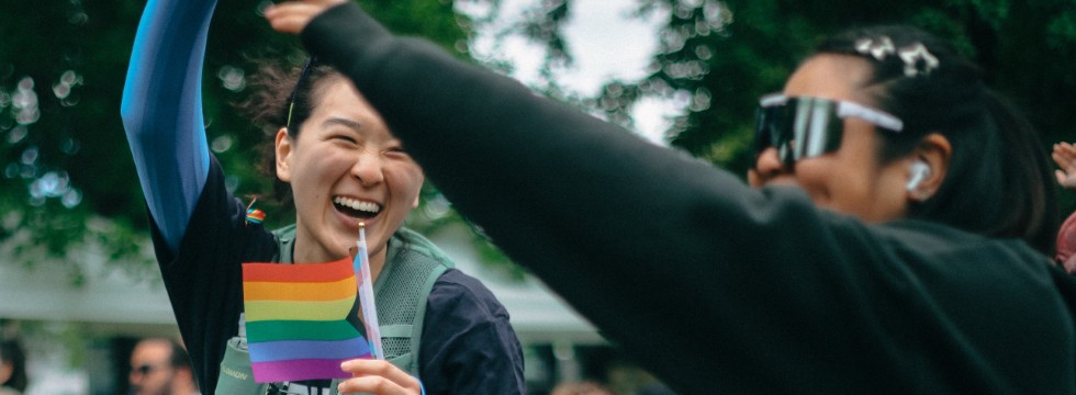 A joyful woman laughs and holds a small Progress Pride flag while high-fiving a friend outdoors.