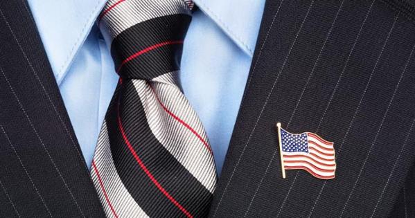 A torso wearing a blue dress shirt and red, black, and silver striped tie underneath a black pinstripe suit jacket with an American flag lapel pin.