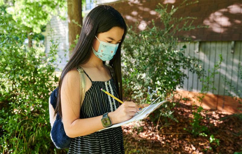 photo of a young person outdoors writing on paper, she is wearing a mask