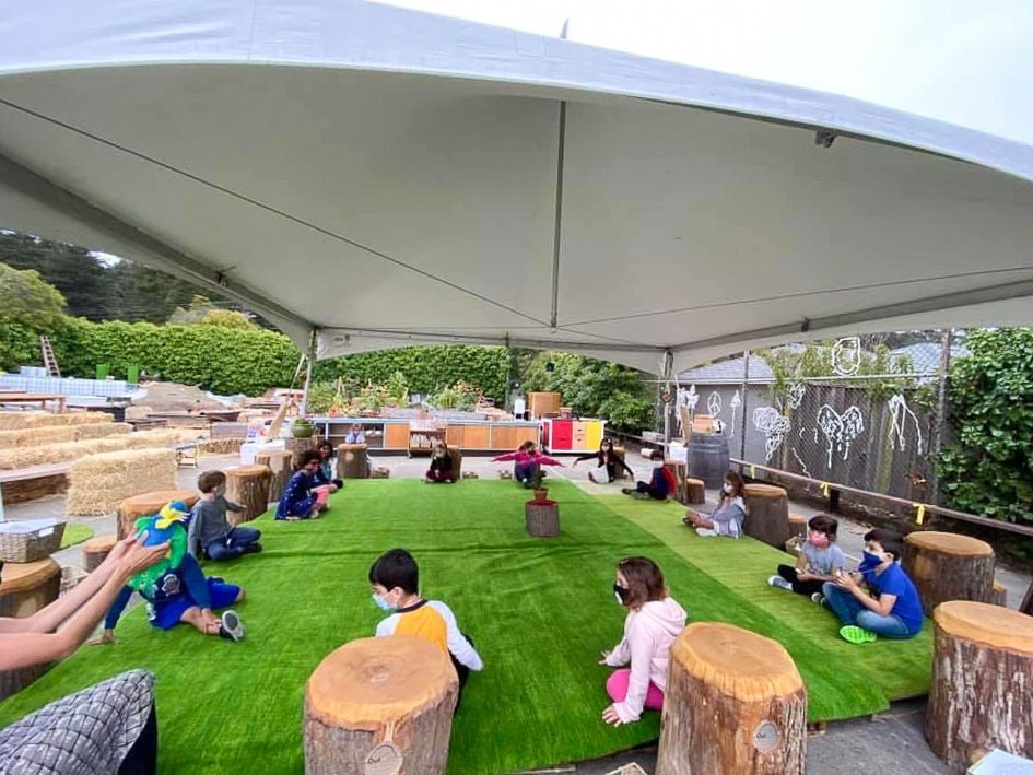 photo of a tented outdoor space, masked children sitting spaced apart
