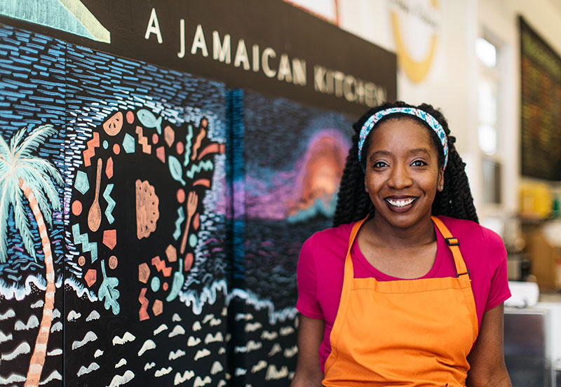 photo of a woman in a chef's apron, in front of a Jamaican restaurant