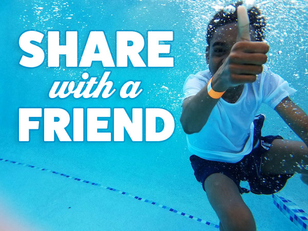 A boy in a white shirt and black swimming trunks gives a thumbs up sign underwater in a swimming pool.