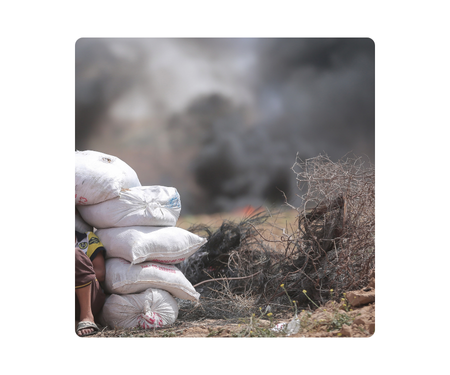 A person crouching behind a stack of white sacks with smoke in the background.