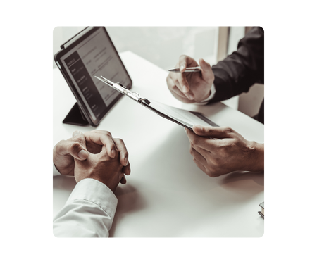 a close-up image of two people's hands around a conference room table discussing a document; a tablet is propped up in the background