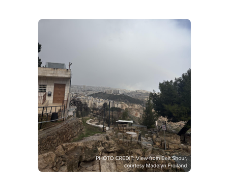 Overcast hillside view with a winding path and city in the distance. Transcribed Text: PHOTO CREDIT: View from Beit Shour, courtesy Madelyn Froiland.