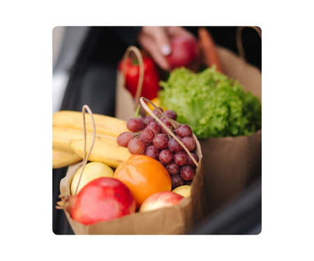 Paper grocery bags filled with fruits and vegetables in a car trunk, including bananas, grapes, lettuce, and more.