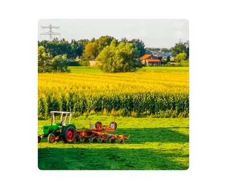 Green tractor on a green field with a cornfield and farmhouse in the background.