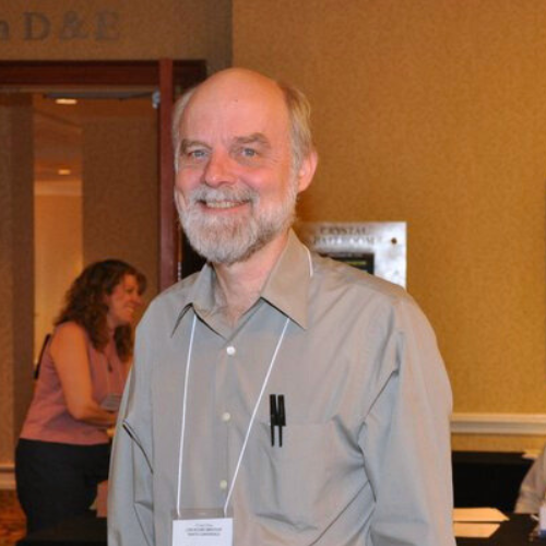 Linton Joaquin wearing a light grey long sleeve button up with two pins in his left chest pocket and a white lanyard tag around his neck smiling at the camera