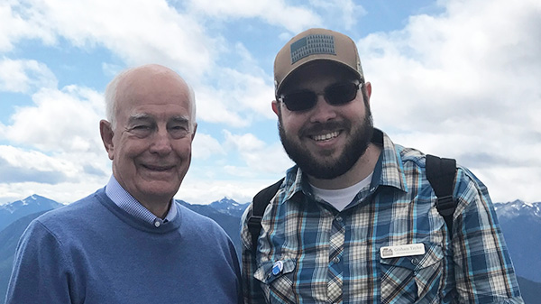 two people stand with mountains and clouds in the background.