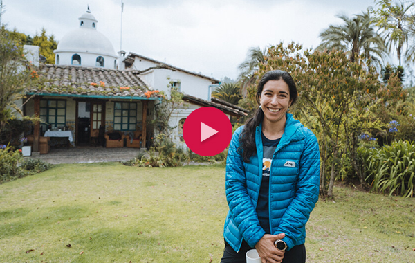 Josy Cardoso smiling in front of a house and greenery with a white video play button placed on top