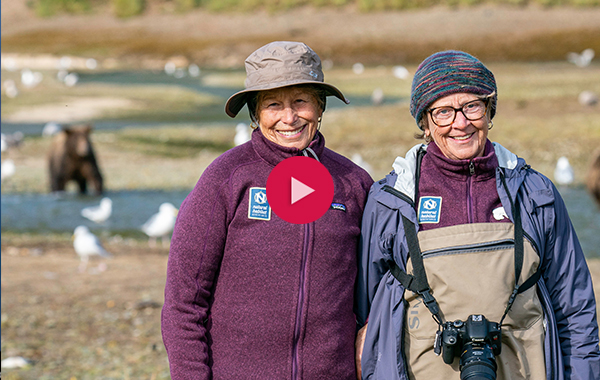 two women posing in front of geyser with a red video play button over the image