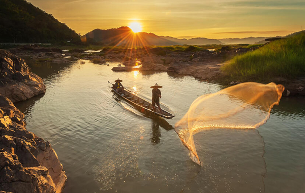 Fisher casts net while on boat with a sun setting behind a mountain in the distance