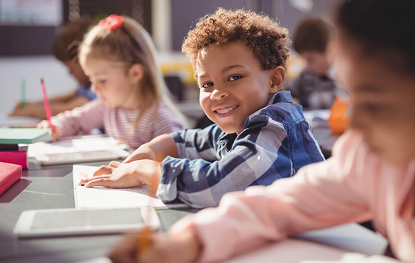 Kids in a classroom sitting at their desks. Three children are visibly pictured with a few in the back that are blurred out. The three children are sitting at desks in a line and the two kids on the outside are slightly blurrier and are writing things in a notebook. The kid in the middle is focused on and is smiling at the camera with hands on a notebook.