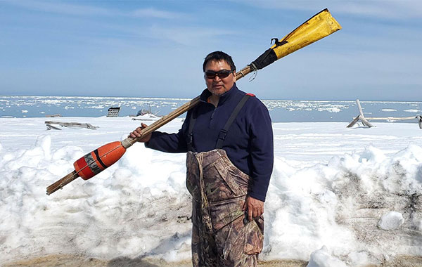 A man wearing sunglasses holds a pole with an orange buoy at one end and a yellow cover at the other in front of the water