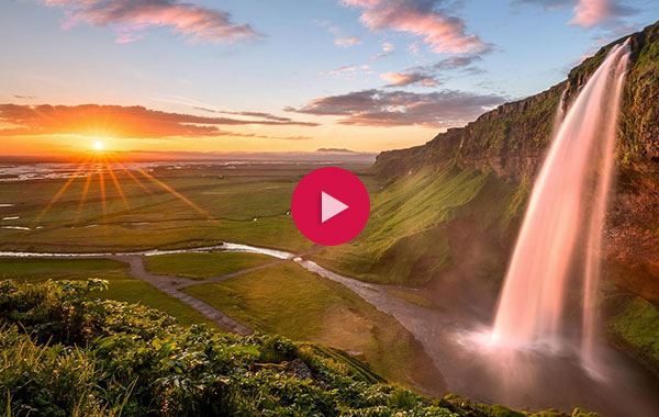 Waterfall in Iceland at sunset. Orange and blue, partly cloudy skies complete the left part of the image and green and grand landscapes with the waterfall make up the right side. A red video play button is overlayed.