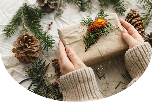 Hands holding stylish paper and twine wrapped gifts with fir branch on rustic table with natural ornaments 