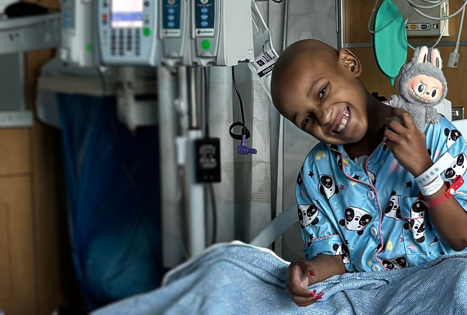young girl with dark skin and no hair smiling while sitting in hospital bed