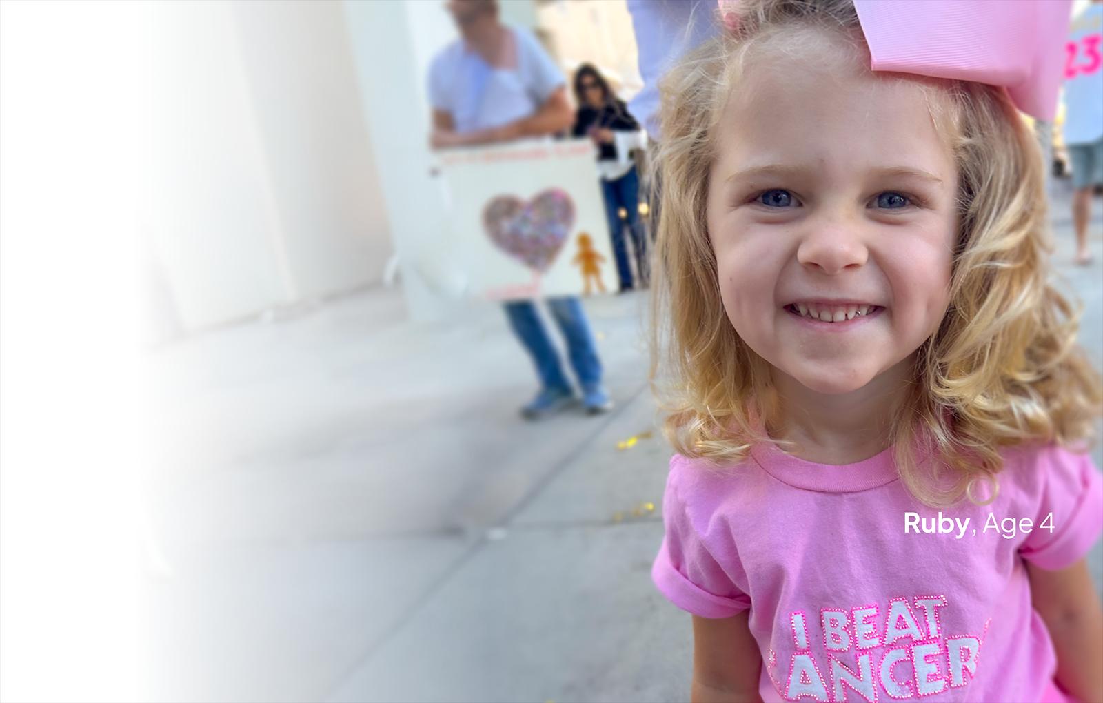 Young girl smiling, wearing pink shirt and bow.