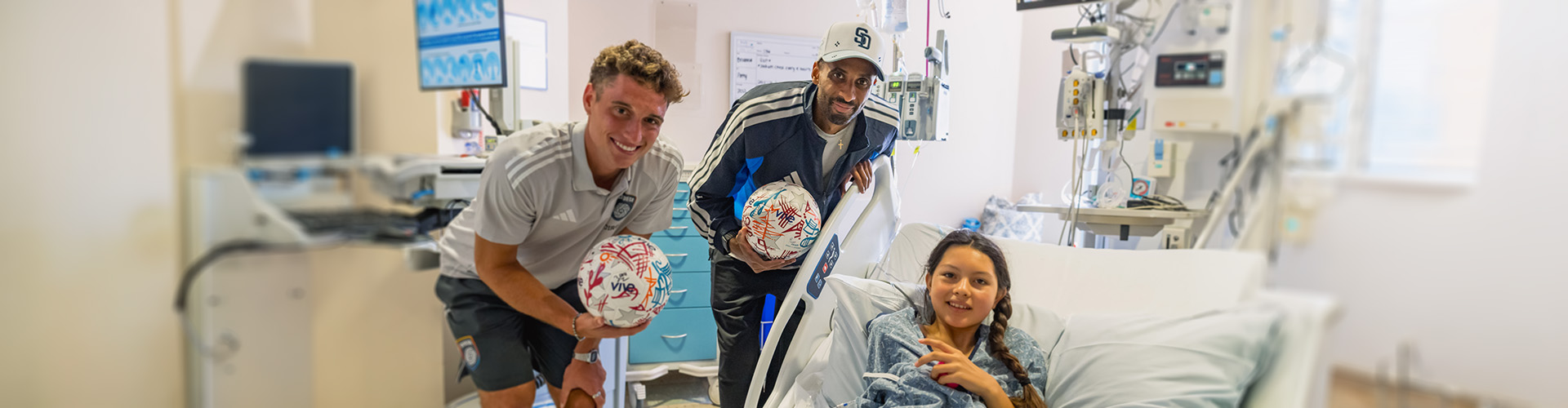Two SDFC players holding soccer balls next to girl in hospital bed