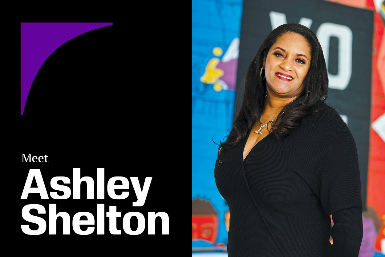 Meet Ashley Shelton. A photo portrait of a Black woman with long, wavy hair and berry lipstick smiling in front of a colorful voting rights-themed banner.