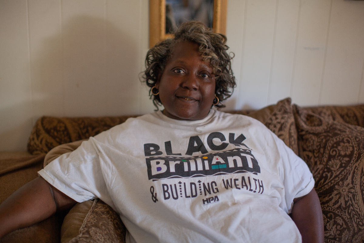 Tina Payne, a Black woman with a soft smile and face-framing salt and pepper curls, is pictured sitting on a sofa.