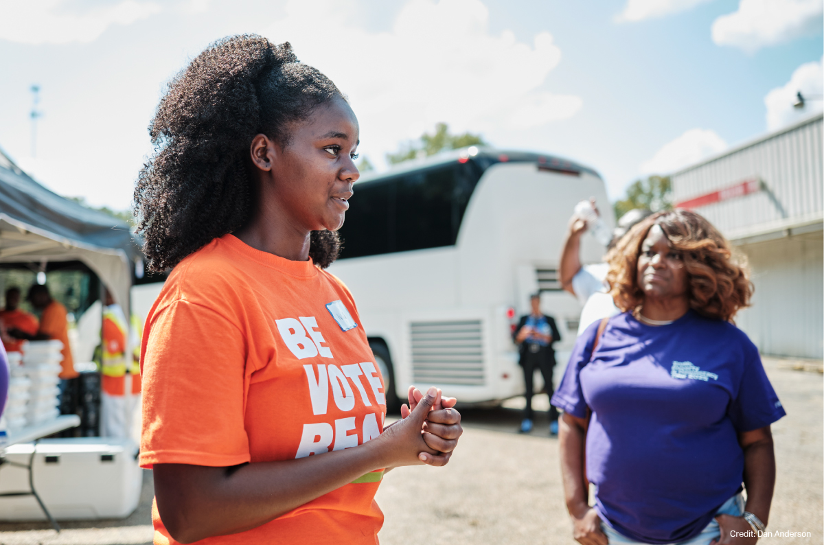 A photo of a pair of Black women in colorful SPLC t-shirts listening intently to off-camera Mississippi residents. A tent, tour bus, and volunteers make up the background.