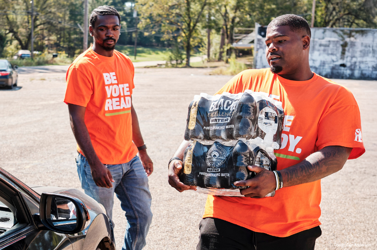 A pair of young Black men in orange SPLC t-shirts bring cases of water to a Mississippi resident waiting in their car. 