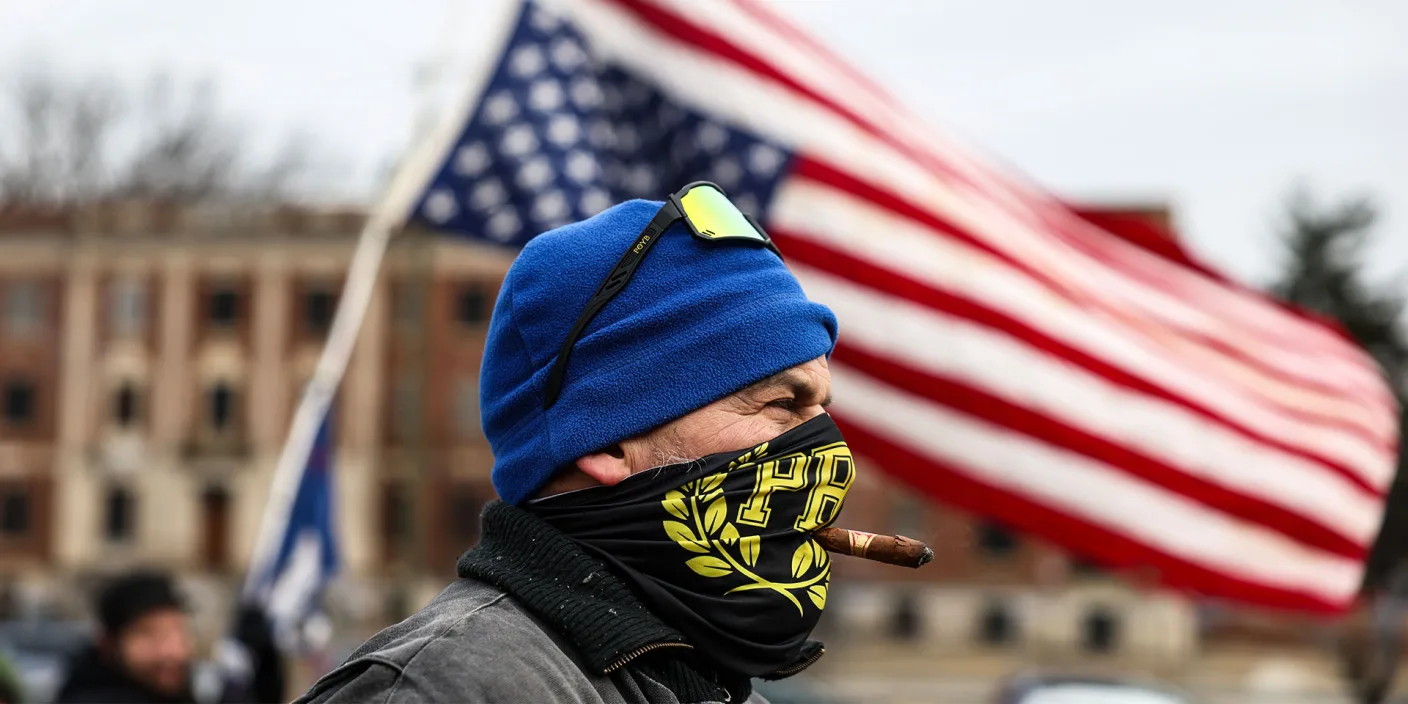 A photo of a white male member of the Proud Boys wearing a bandana over his face and smoking a cigar on Jan. 6. An upside-down U.S. flag flies behind his head.