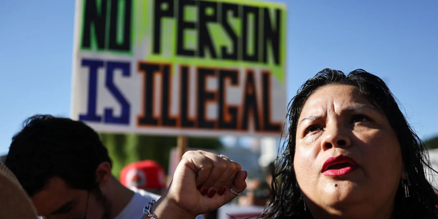 A photo of a Brown woman raising her fist while gazing towards the sky at a protest. A sign that reads 