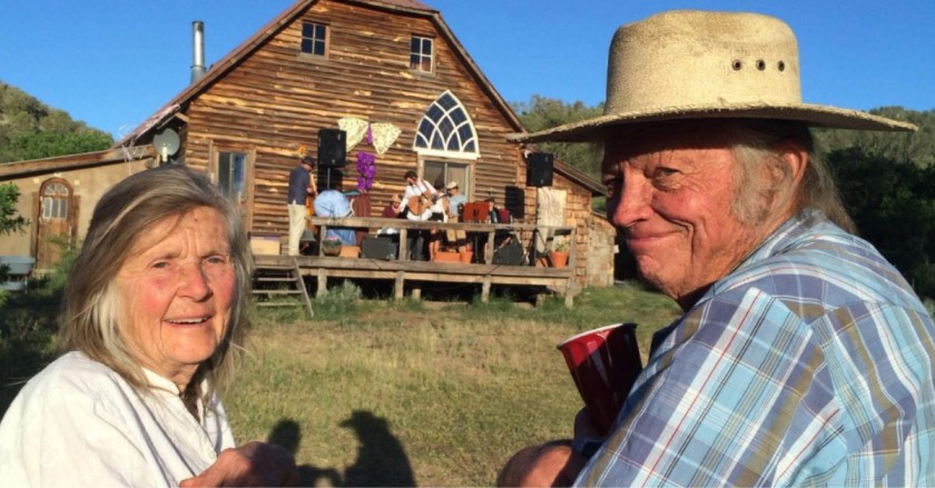 Smiling older couple outdoors near a rustic wooden building, with a small live band performing on a porch behind them.