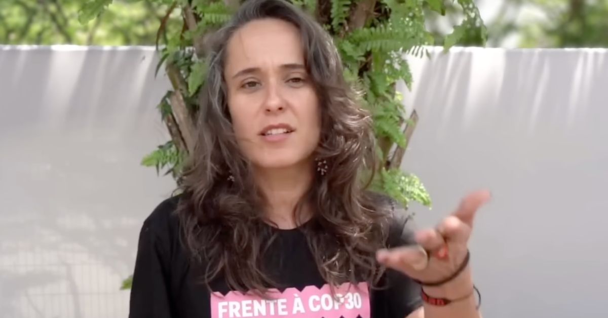 A woman speaking to the camera with plants and a wall behind her.