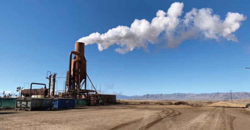 Industrial facility venting thick white smoke into the air under a clear blue sky.