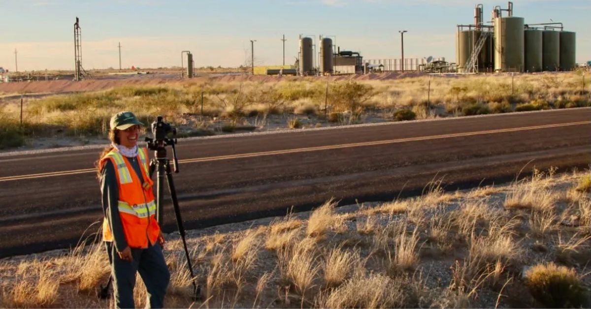 A person in a safety vest records an oil and gas facility across a rural road, documenting industrial activity in the landscape.