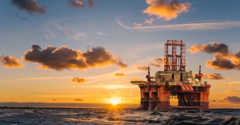 Offshore oil platform in open water at sunset, silhouetted against orange sky and scattered clouds.
