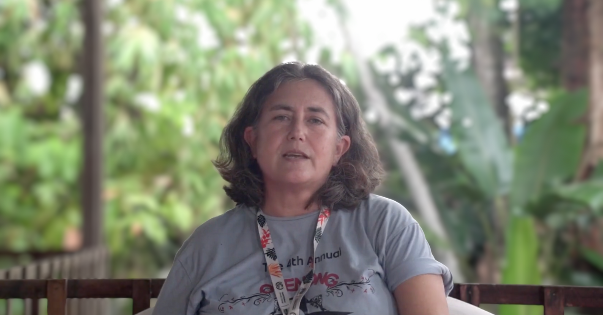 A woman speaking on camera outdoors with blurred greenery behind her.